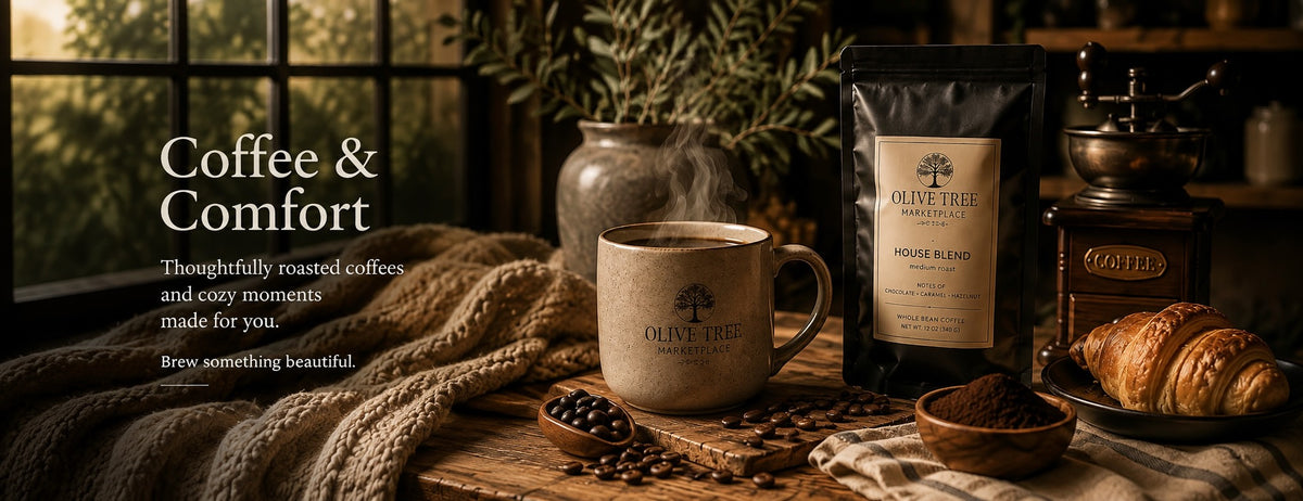 Olive Tree Marketplace coffee and comfort — a steaming stoneware mug, House Blend coffee bag, vintage grinder, croissants and coffee beans on a wooden table with a knitted throw