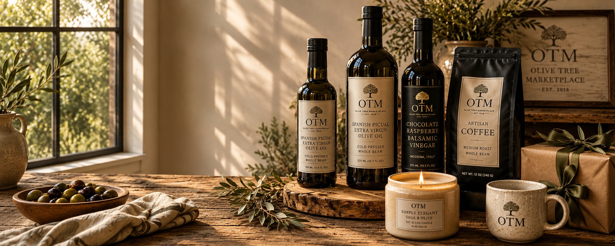 Olive Tree Marketplace olive oils, balsamic vinegar, artisan coffee, a lit candle, a bowl of olives and a wrapped gift arranged on a rustic wooden table in warm window light